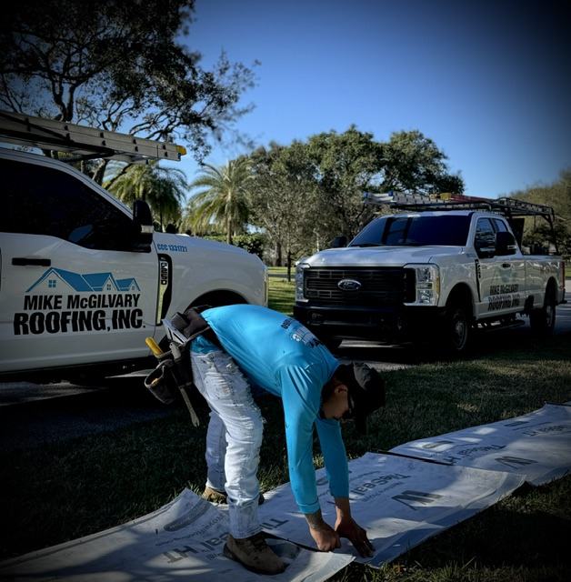 A Mike McGilvary Roofing crew member preparing underlayment materials beside two branded company trucks, license CCC1331721 visible