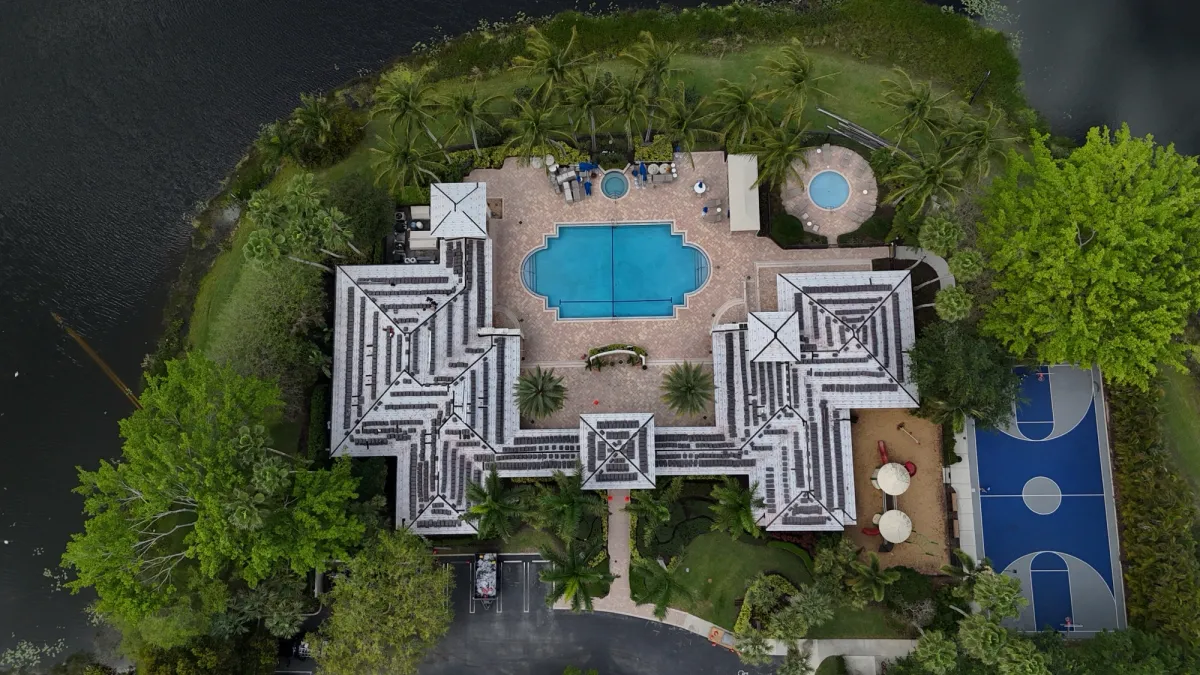 Drone view of a lakefront HOA clubhouse with stacks of new roof tile loaded across a fully underlaid roof, next to a pool and basketball court