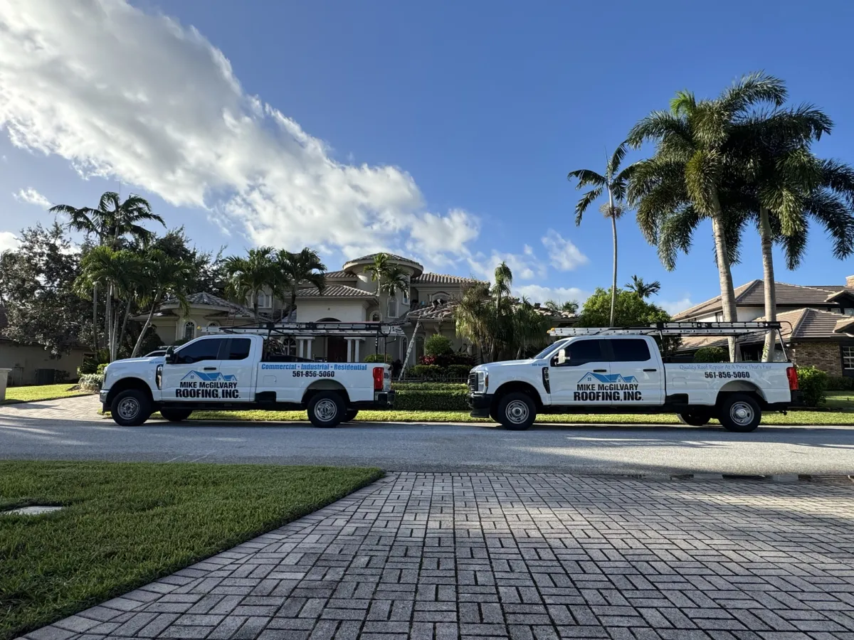 Two white Mike McGilvary Roofing trucks parked in front of a tile-roof home with palm trees.
