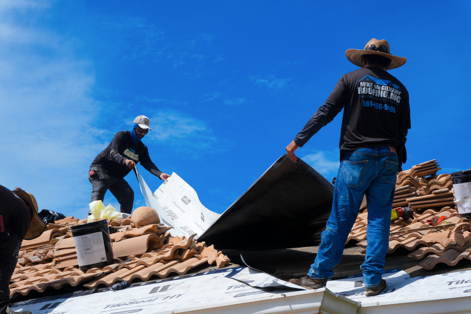 Two Mike McGilvary Roofing crew members rolling out new underlayment on an opened section of a tile roof