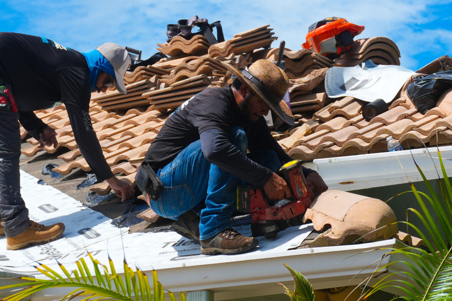 Roofers fastening new underlayment with a nail gun, with the removed roof tiles stacked on the roof beside them